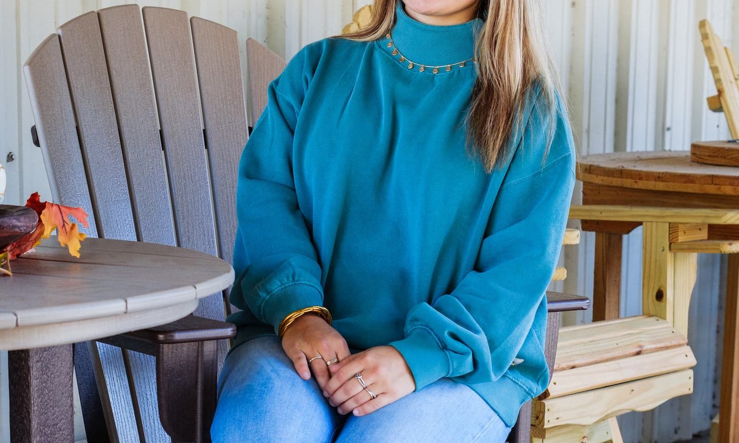 Woman sitting on a tall outdoor chair at Anderson’s General Store, wearing a teal pullover, light-wash jeans, and brown boots, smiling in a cozy fall setting.