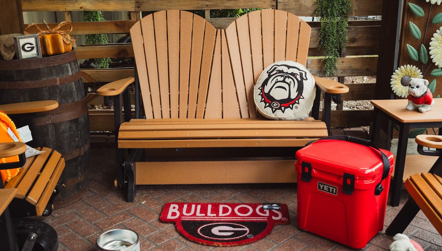 Outdoor patio setup with Georgia Bulldogs-themed decor, including a red YETI cooler, Bulldogs mat, dog bowl, and cushion, surrounded by wooden furniture.