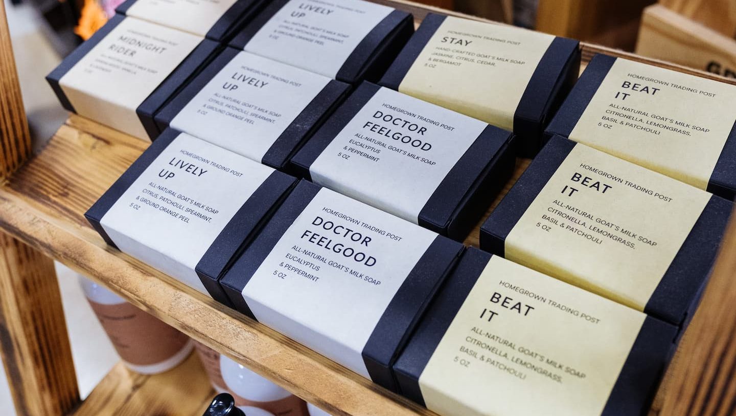 Display of handcrafted goat’s milk soaps from Homegrown Trading Post at Anderson’s General Store, featuring scents like Doctor Feelgood, Beat It, and Lively Up arranged neatly on wooden shelves.