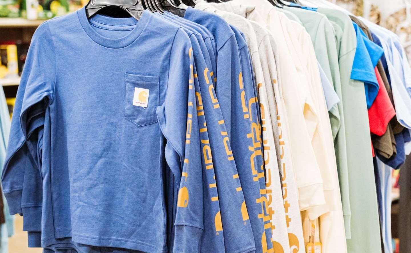 Rack of kids’ Carhartt long-sleeve shirts in blue, gray, cream, and green tones displayed neatly on hangers inside Anderson’s General Store.