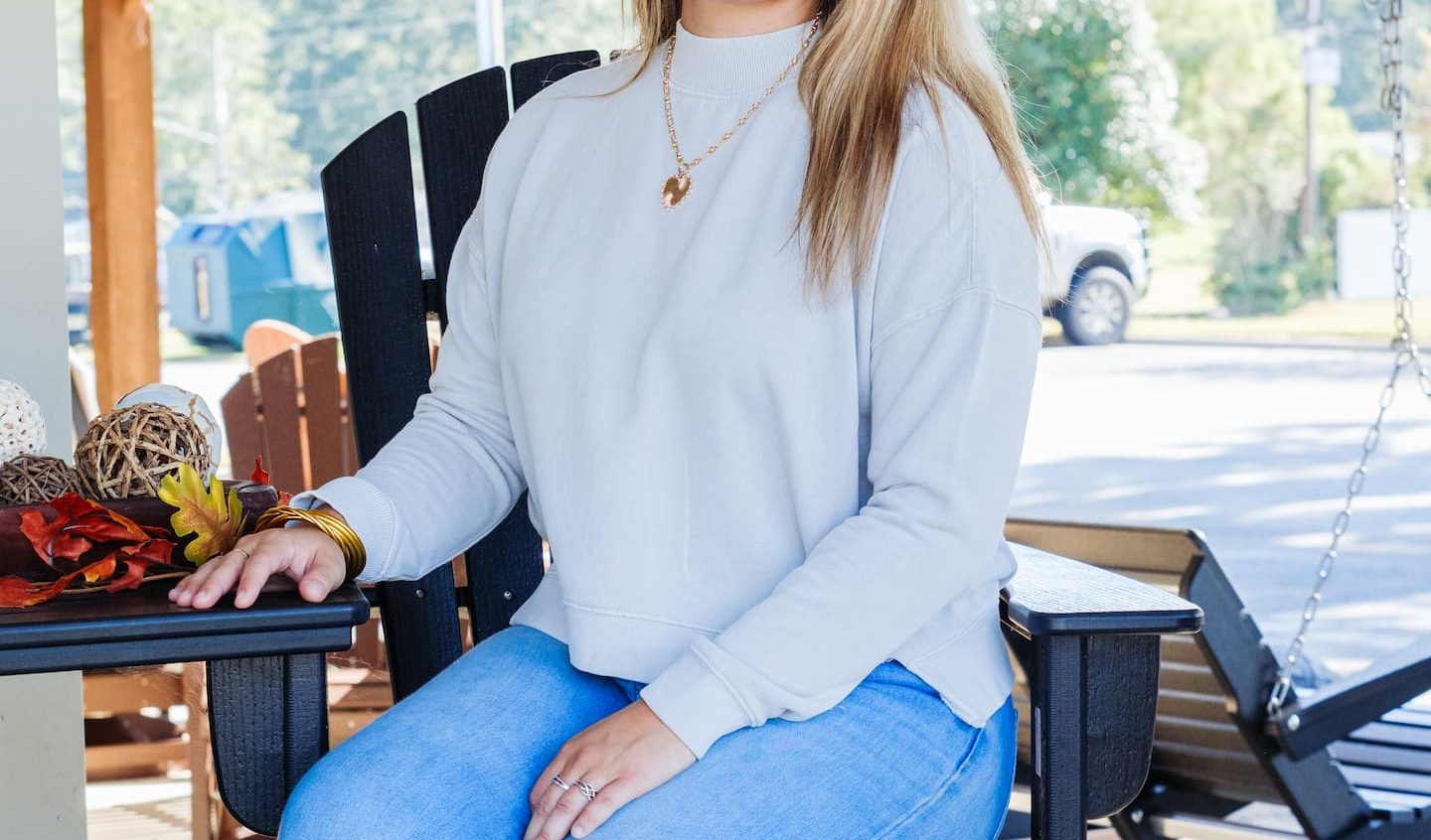 Woman sitting on a tall outdoor chair at Anderson’s General Store wearing a light gray pullover, light-wash jeans, and brown boots, smiling in a casual fall outfit.