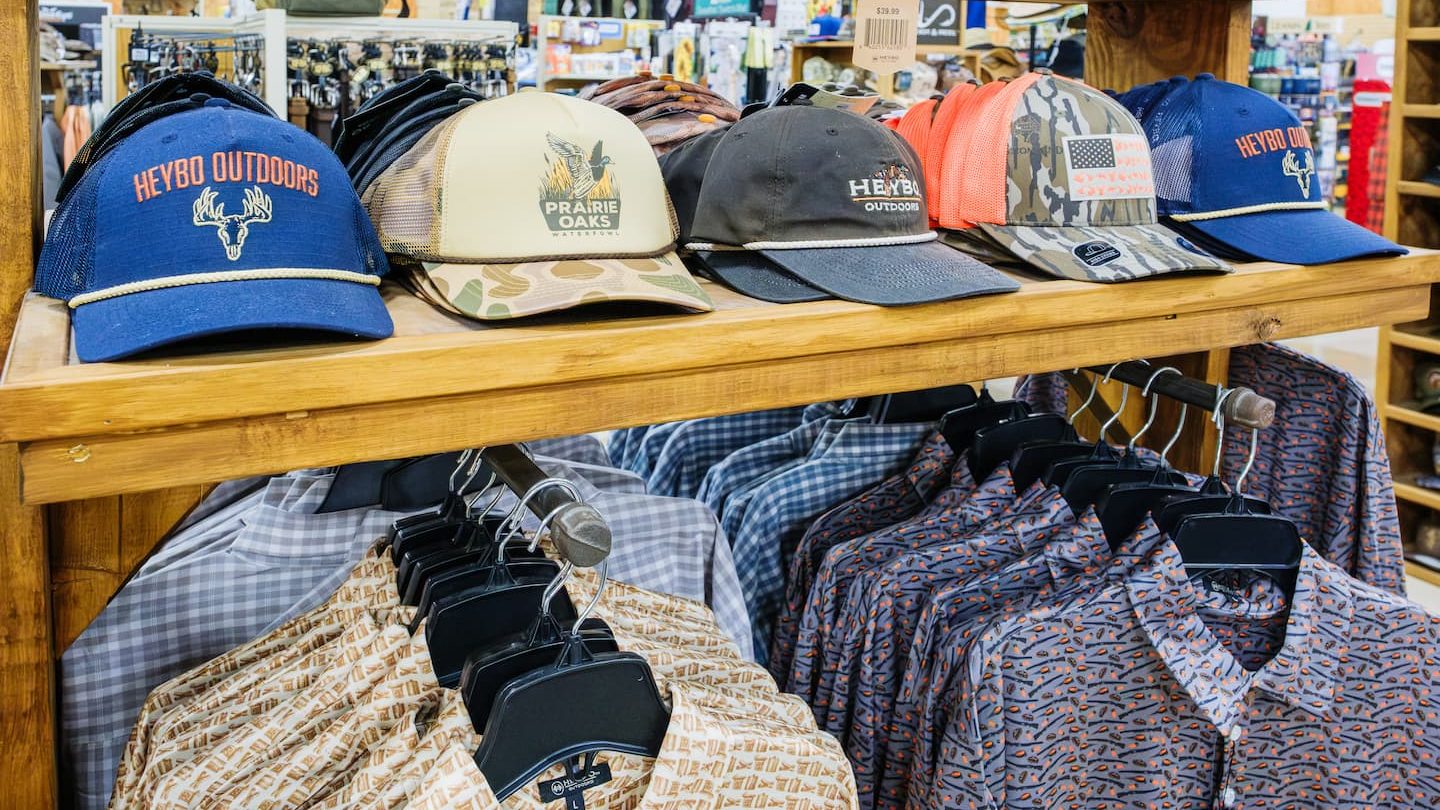 Display of Heybo Outdoors and Prairie Oaks hats with camo and American flag patch designs, paired with patterned button-down shirts at Anderson’s General Store — featuring leather patch hats, mesh back outdoor hats, and southern hunting apparel.