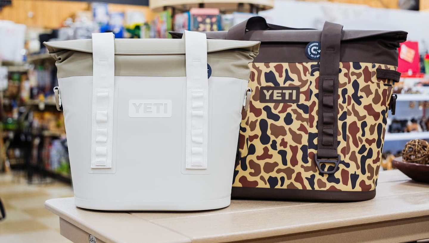 Two YETI soft coolers on display at Anderson’s General Store — one in solid white and one in a camo pattern — featuring durable straps and leak-resistant designs perfect for outdoor adventures.