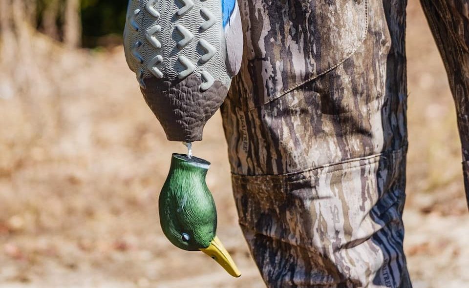 Hunter dressed in camouflage holding a duck decoy by the neck, standing outdoors during hunting season.