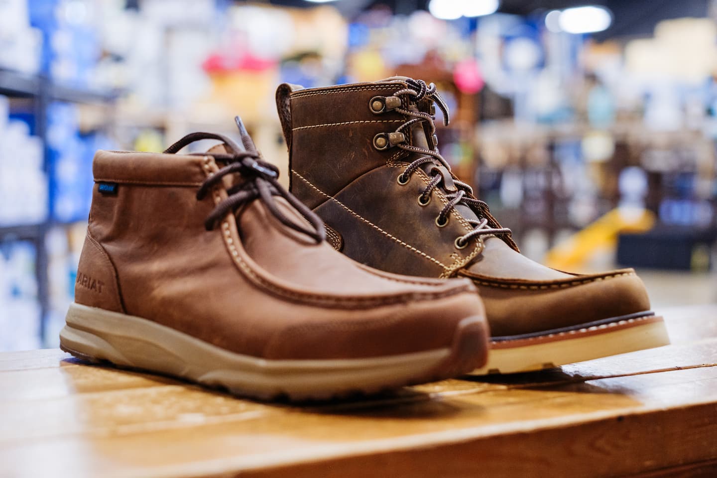 Close-up of two pairs of brown Ariat work boots displayed on a wooden table — one ankle-height casual style and one rugged lace-up design, available at Anderson’s General Store.