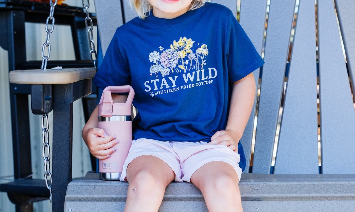 Smiling child sitting on a porch swing wearing a navy ‘Stay Wild’ Southern Fried Cotton T-shirt, pink shorts, and white shoes, holding a pink YETI bottle at Anderson’s General Store.