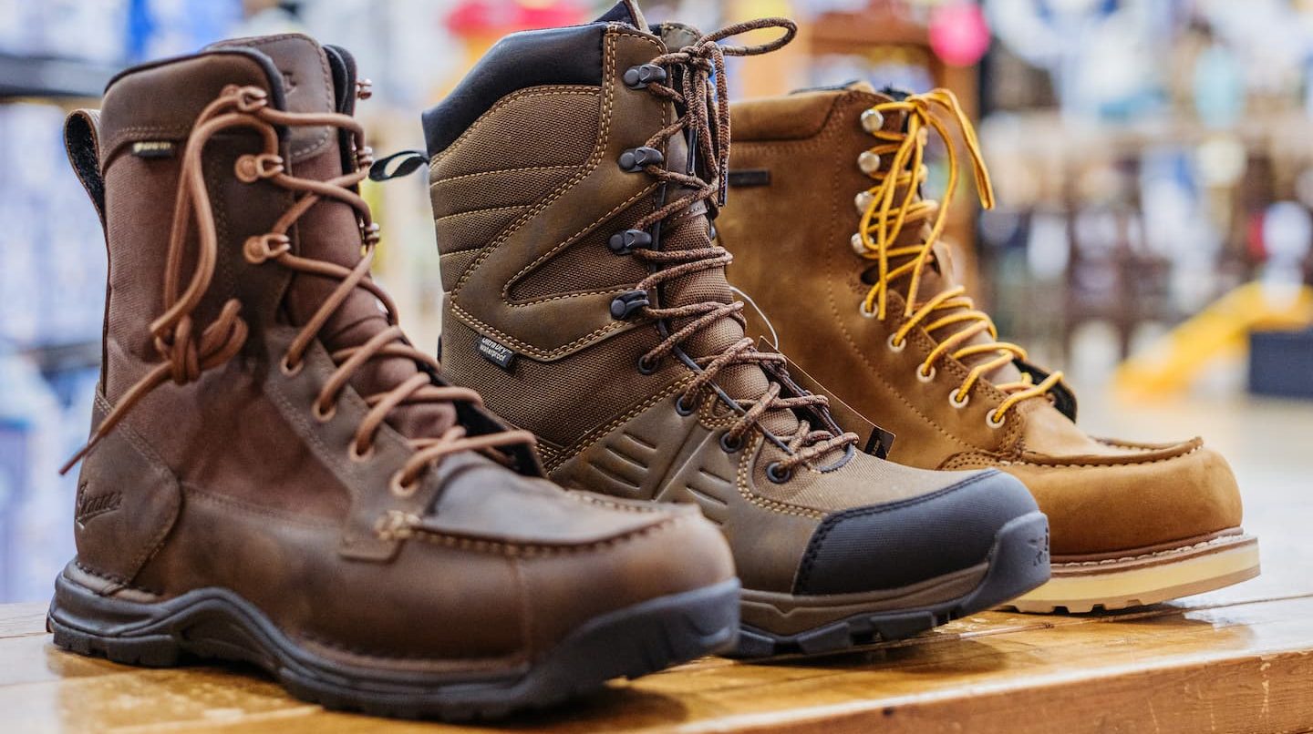 Close-up of rugged work and hiking boots in brown and tan tones displayed on a wooden table — durable outdoor footwear available at Anderson’s General Store.