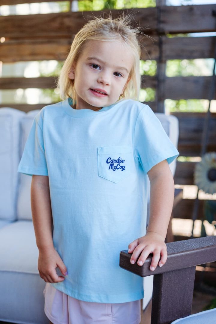 Young child wearing a light blue Cardin McCoy T-shirt with a front pocket, standing outdoors next to patio furniture with a wooden fence and greenery in the background.