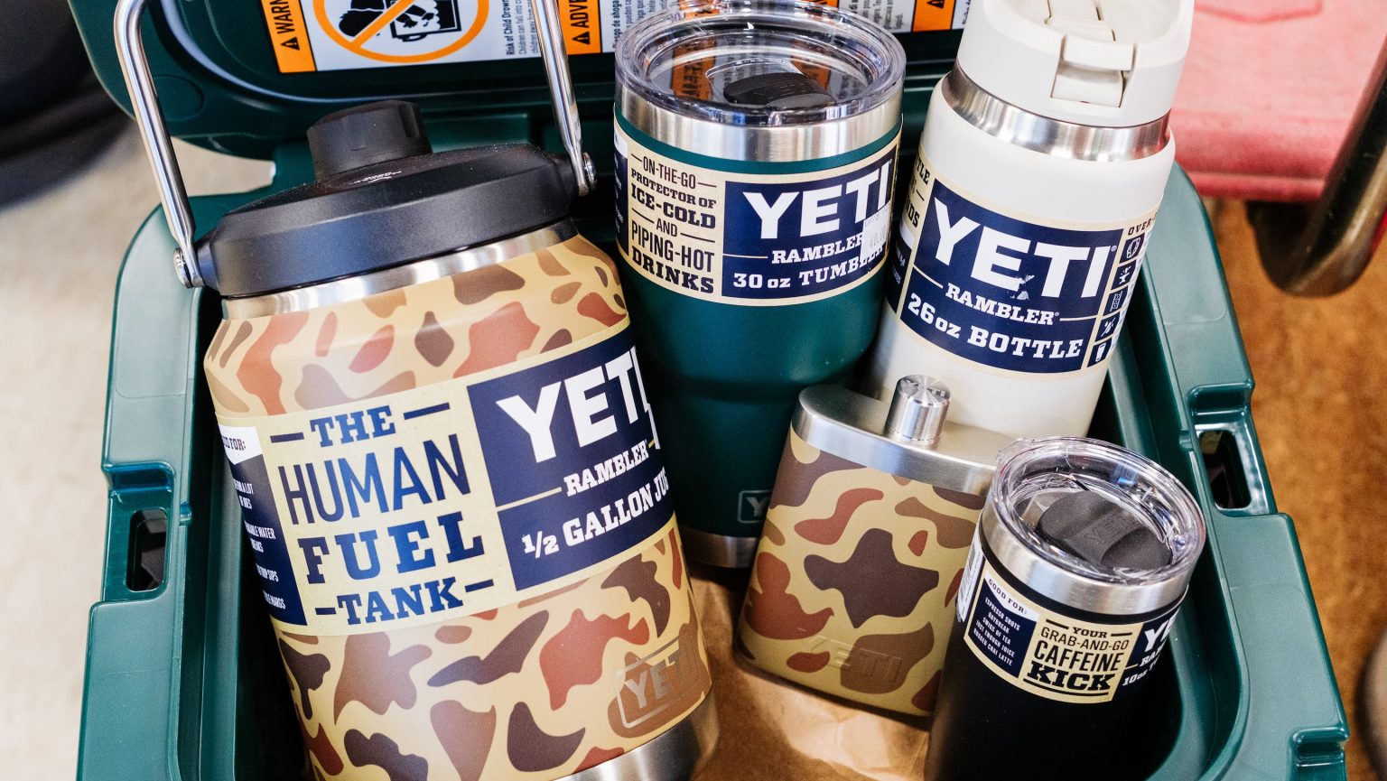Assortment of YETI drinkware in a green cooler, including camo-patterned and solid color Ramblers, bottles, and jugs labeled with fun phrases like ‘The Human Fuel Tank’ — durable stainless-steel drinkware perfect for gifts at Anderson’s General Store.