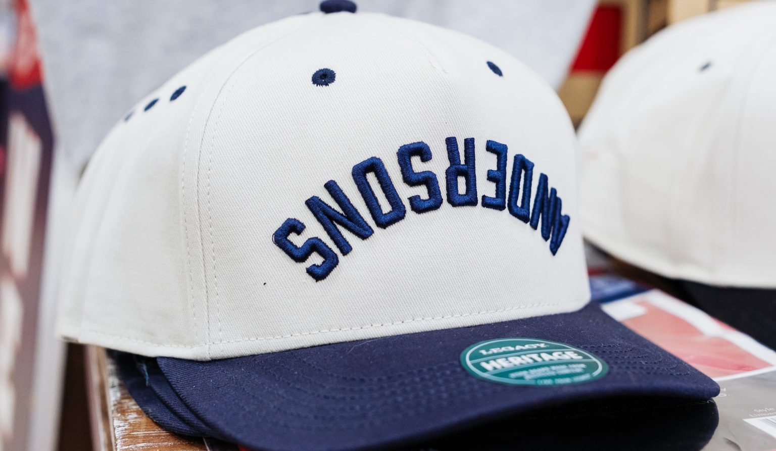 Close-up of a white and navy baseball cap embroidered with the word ‘Anderson’s’ in blue, displayed on a wooden counter at Anderson’s General Store.