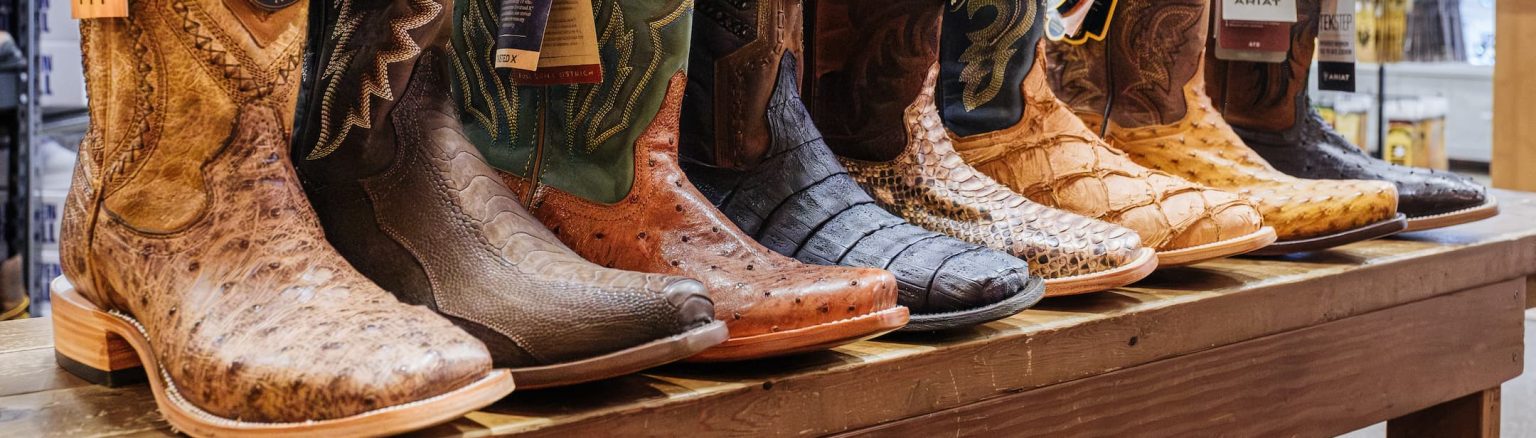 Row of cowboy boots from brands like Corral, Dan Post, and Ariat displayed on a wooden table — premium western boots in various colors and exotic leathers available at Anderson’s General Store