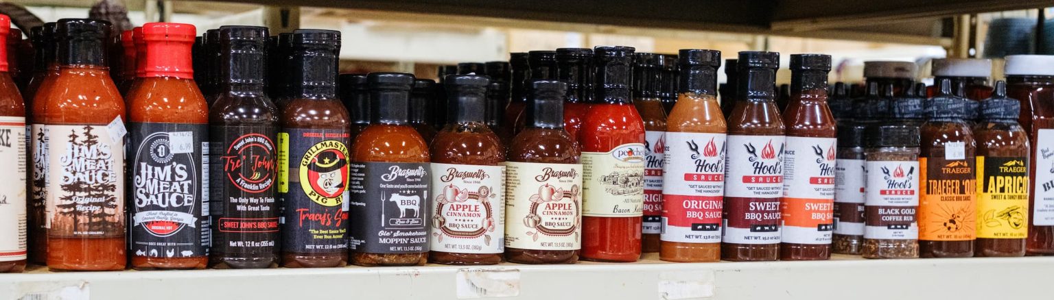 Assorted BBQ sauces, rubs, and seasonings from brands like Traeger, Bulloch, Hoot’s, and Busha’s displayed on shelves at Anderson’s General Store.