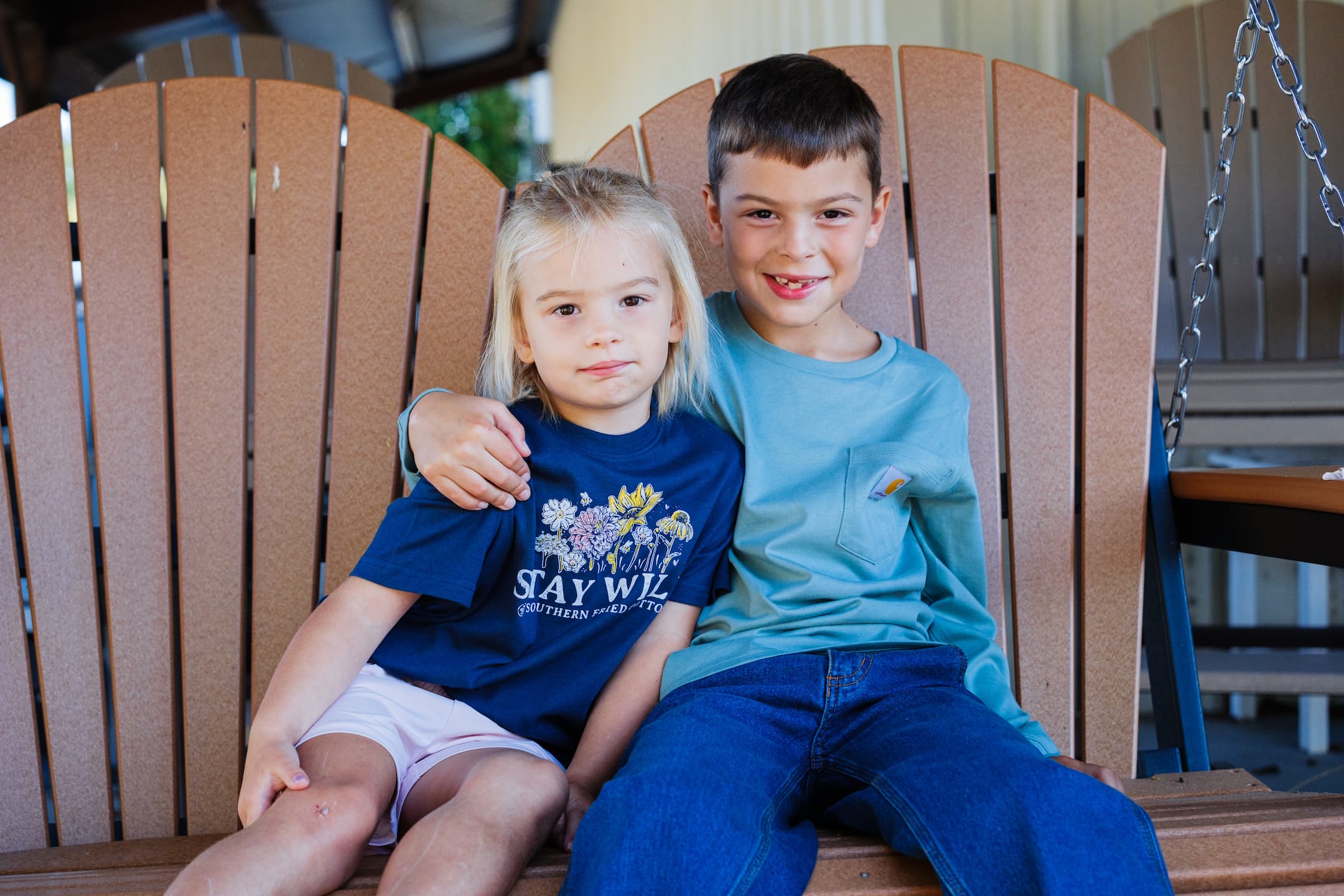 Two smiling kids sitting together on a porch swing at Anderson’s General Store, wearing casual Southern Fried Cotton and Carhartt shirts, enjoying a cozy outdoor moment.