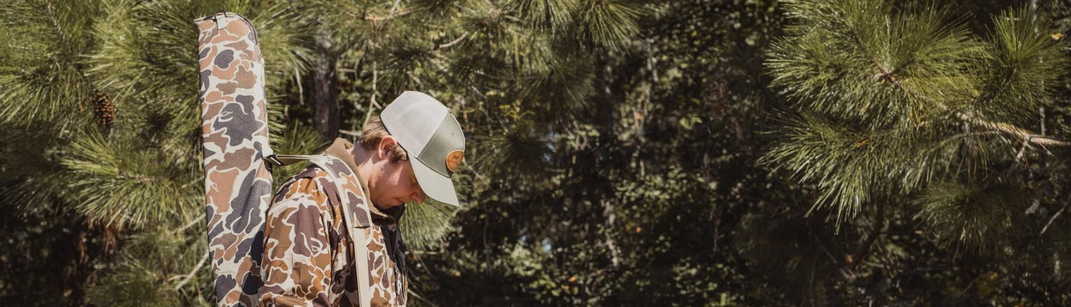 Hunter dressed in camo jacket and khaki pants walking through the woods with a Drake Waterfowl gun case slung over his shoulder, surrounded by pine trees in South Georgia.