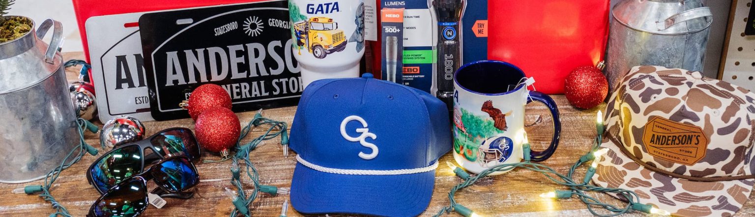 Holiday gift display from Anderson’s General Store featuring wrapped presents, Georgia Southern gear, Anderson’s license plates, a NEBO flashlight, Costa sunglasses, mugs, tumblers, and festive lights and ornaments on a rustic wooden table.